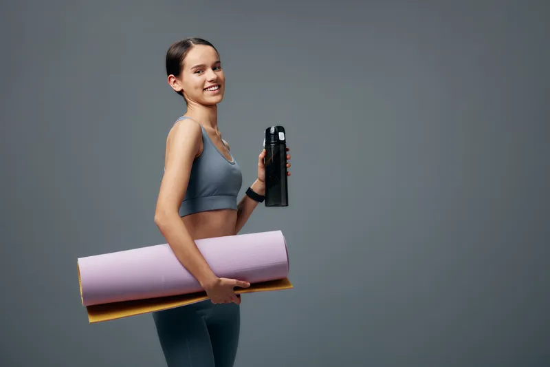 Fitness woman in athletic wear stands with a water bottle and yoga mats against a plain backdrop, ready for training, warm up and an efficient workout session in the gym.