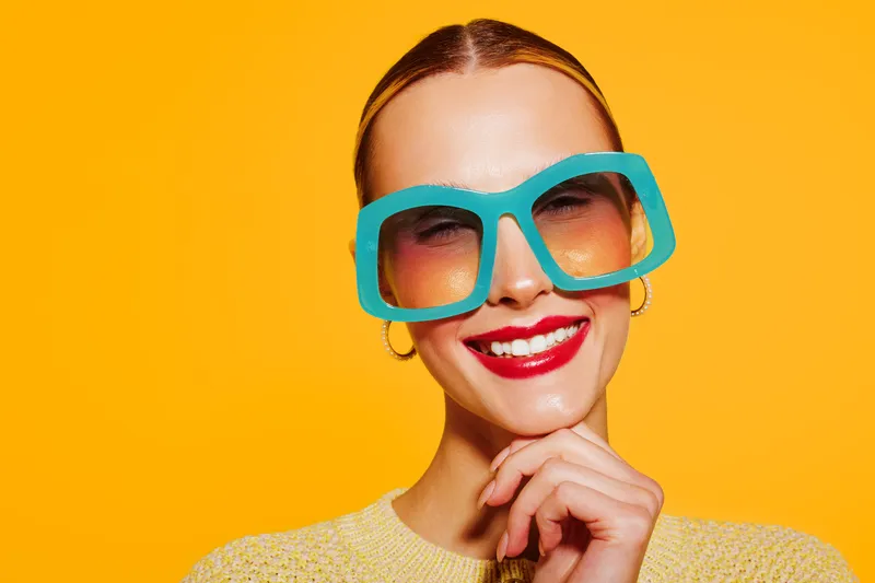 Smiling woman with bright red lipstick wears oversized turquoise sunglasses and yellow sweater. Vibrant orange background creates cheerful and playful mood in this close-up portrait.