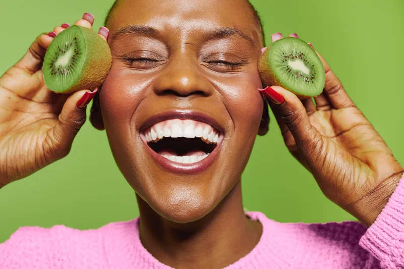 Happy African woman with dark skin holds two kiwi halves near her face, smiling with closed eyes. Bright green background and pink sweater create a vibrant fresh concept of healthy lifestyle.