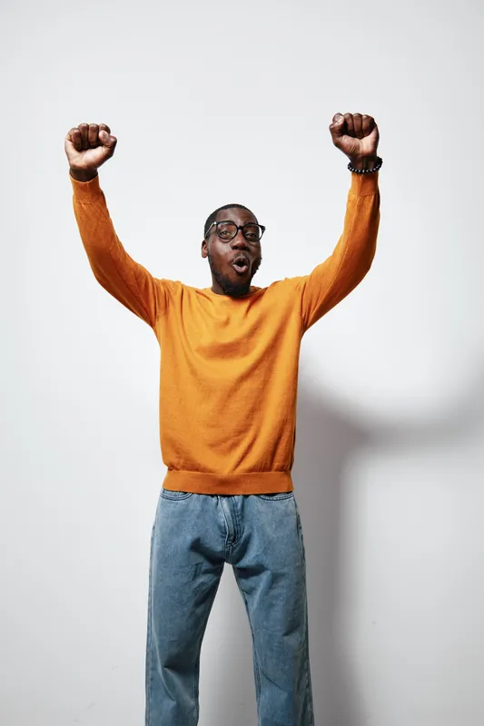 African American man with dark skin wearing orange sweater and blue jeans celebrating with raised fists and joyful expression against white background in studio.