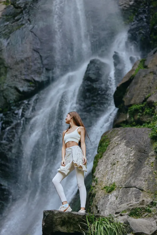 Woman standing near waterfall in activewear, surrounded by natural rocks and greenery, showcasing strength and serenity in outdoor adventure setting.
