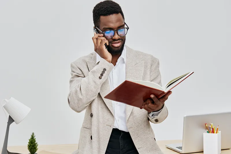 Confident professional man talking on mobile phone while reading a notebook in bright modern office space, wearing smart casual beige blazer and glasses, working or communicating remotely, business li