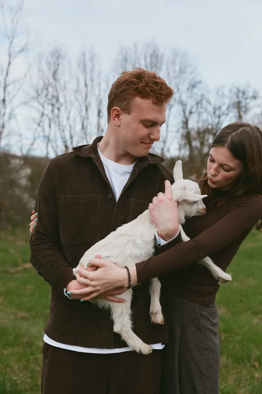 Caring couple outdoors hugs a playful goat kid, showcasing lifestyle, affection, and nature connection. Tender moment of trust, joy, and companionship in daylight.