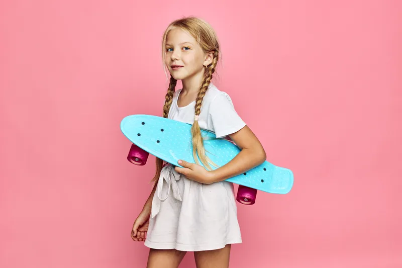 Blonde girl with braided hair holding a blue skateboard against a pink background. Casual clothing and confident expression. Concept for youth and leisure.