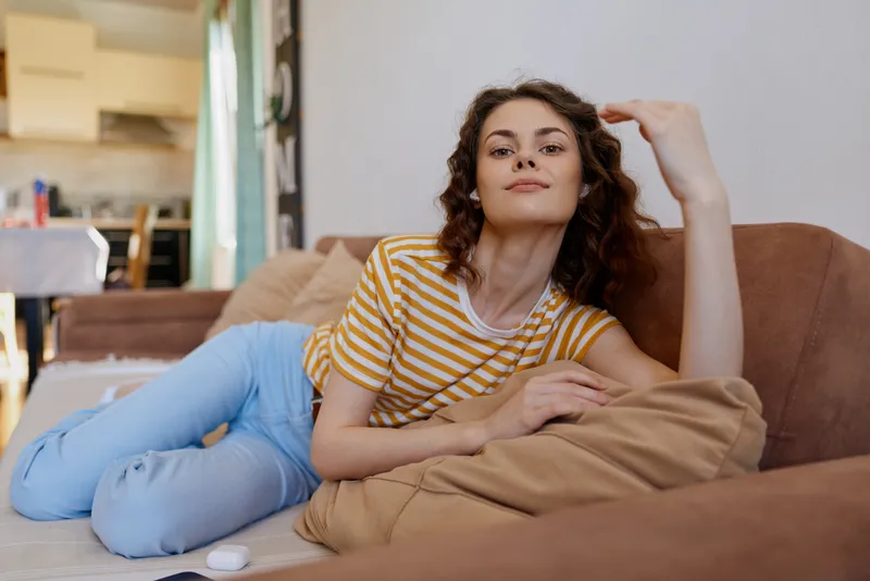 Relaxed woman lounging on a couch at home, wearing a striped tee and light jeans. Soft natural light highlights a calm relaxation moment, suggesting comfort and everyday self care daily pose.