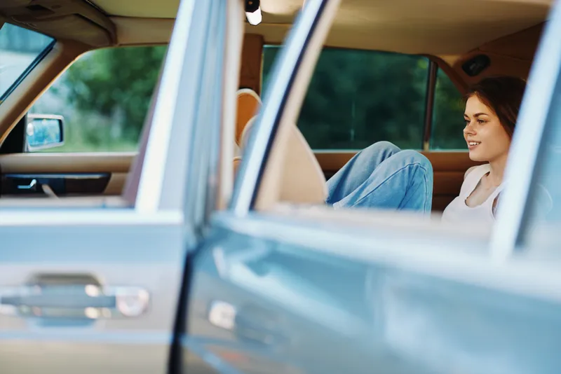 Young woman relaxing in a vintage car with her legs up, enjoying a sunny day outdoors, showcasing casual fashion and a carefree spirit.