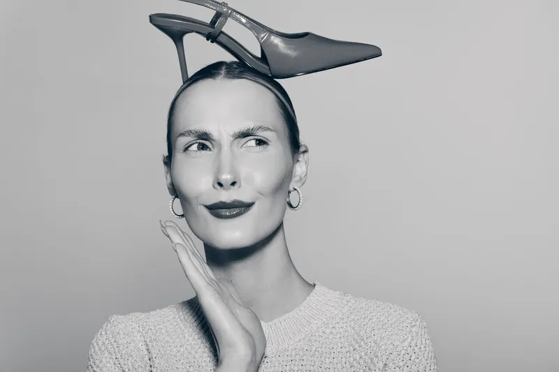 Caucasian woman with fair skin wearing a high heel shoe on her head looks confused isolated on grey background. Concept for fashion, humor, and irony.