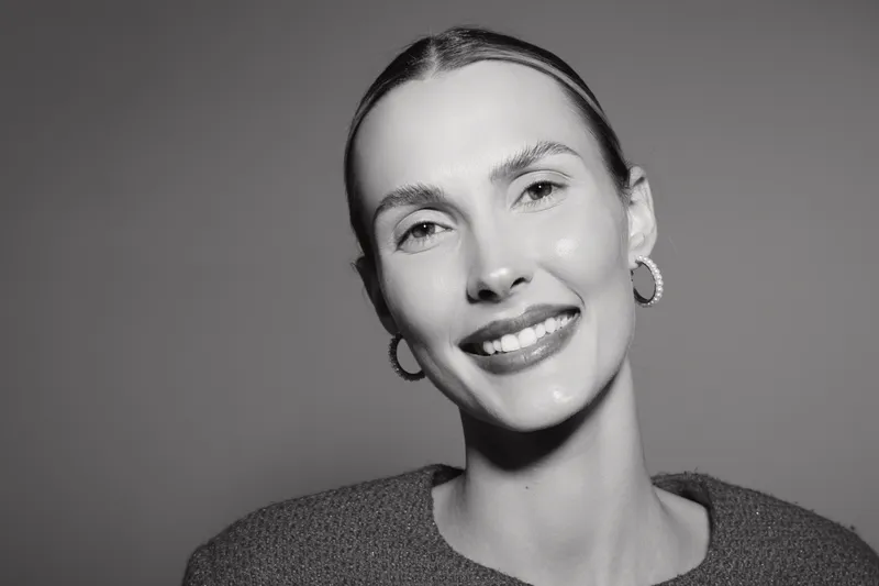Smiling white woman with light skin and slicked back hair poses against grey background wearing hoop earrings and knit sweater. Portrait concept of happiness and confidence.