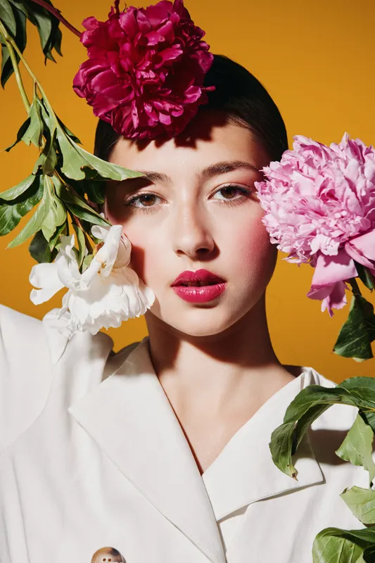 Young woman with bright makeup poses with fresh pink, white, and red flowers framing her face on a vibrant orange background. Concept of beauty, fashion, and creative portrait.