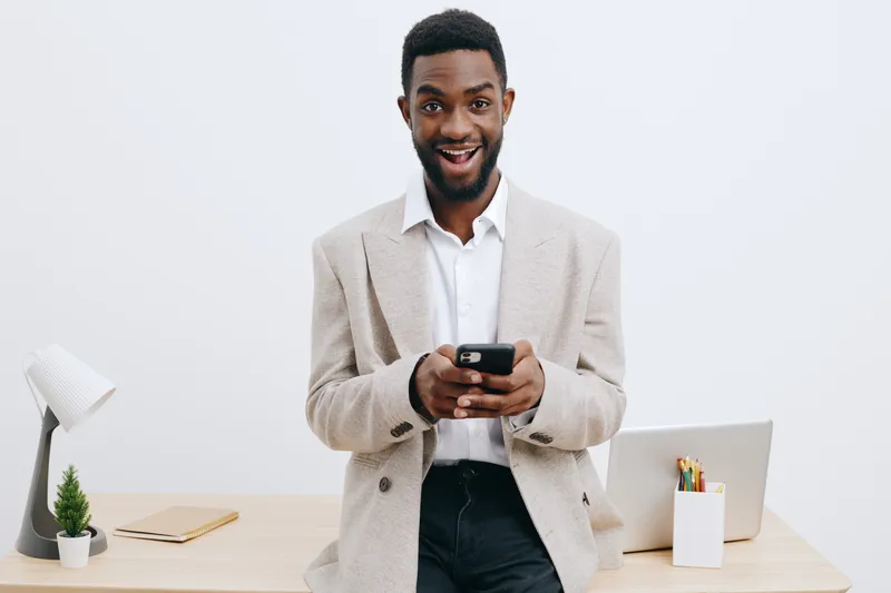 Smiling man in smart casual attire using mobile phone while standing at modern office desk with laptop, plant, and office supplies. Studio portrait of cheerful businessman, technology, communication, 