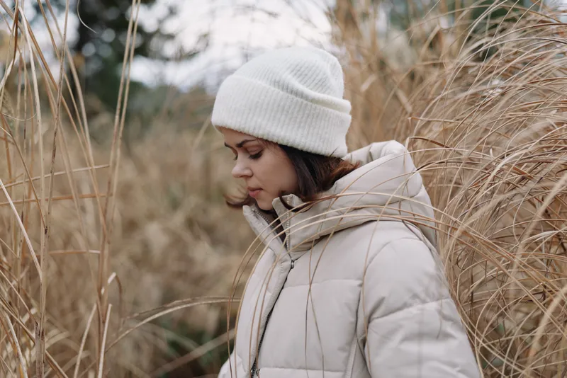 Winter woman in a white puffer jacket and knit beanie stands among tall grasses, capturing a calm outdoor scene with soft textures and muted light.