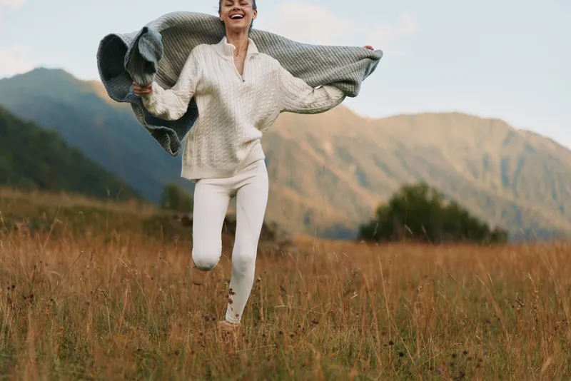 Young woman in white sweater and pants joyfully runs through a grassy field with a blanket in her hands during a clear day in the mountains.