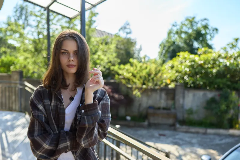 A woman stands on a sunlit balcony in casual plaid clothing, posing for a natural outdoor portrait with a relaxed lifestyle vibe