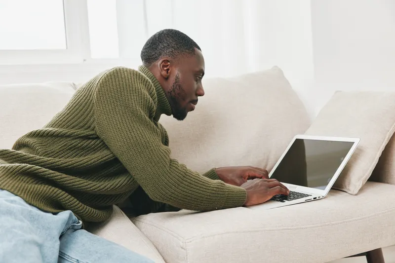 A focused young African American man working on a laptop at home, wearing a cozy green sweater, embodying a sense of productivity and concentration in a bright living space