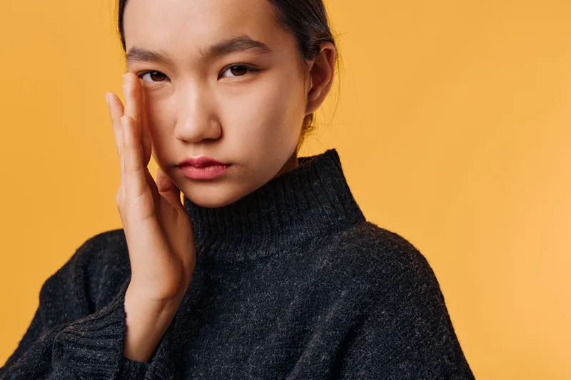Woman with worried facial expression touching face in casual sweater against orange background. Portrait of thoughtful female with hand near cheek showing concern or discomfort.