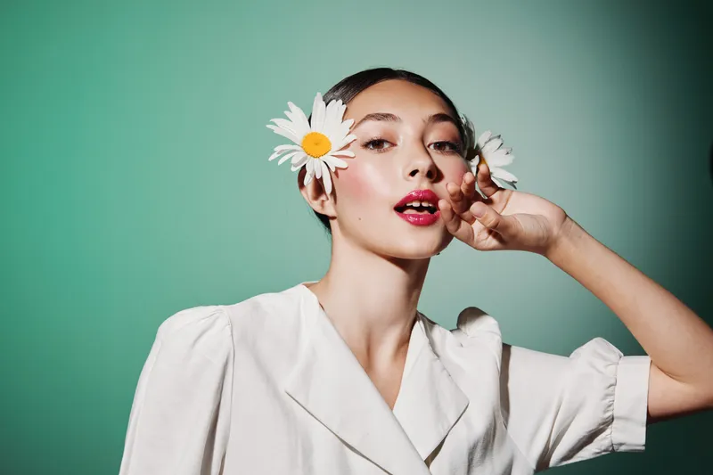 Young woman with clear skin wears white clothing and daisy flowers in her short hair against a stylish green background. Concept of beauty creative fashion and fresh elegance.