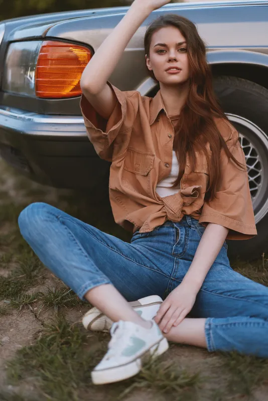 Stylish young woman in casual attire, relaxing beside a vintage car, exuding confidence and a laid-back vibe in a summer setting perfect for lifestyle photography.