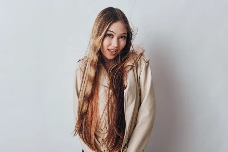 Young woman with long hair wearing beige hoodie posing on white background, smiling and looking at camera for fashion and portrait photography.