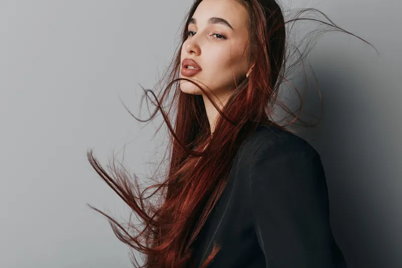 Woman with long red hair and closed eyes posing in black jacket against gray background. Fashion portrait with natural makeup and flowing hair in studio lighting.