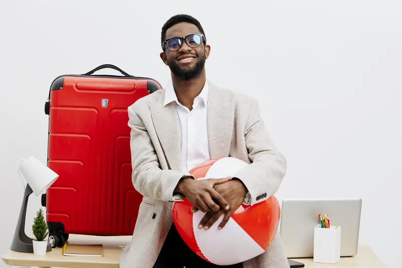Smiling man with dark skin, wearing glasses and a light-colored blazer, holding a red and white paper heart, standing in a bright modern office with a red suitcase, laptop, and plants, celebrating lov