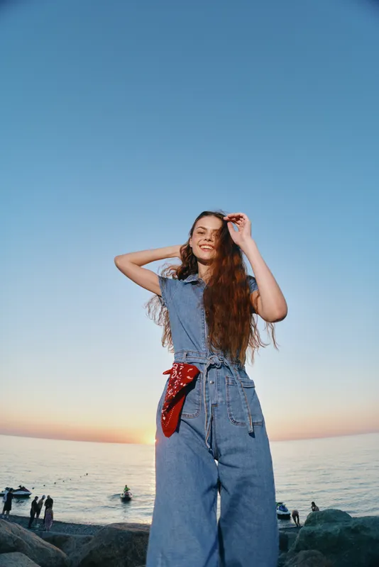 Lifestyle woman in modern boho-western denim street style enjoying sunset by the sea, with film color warmth and relaxed smiling expression