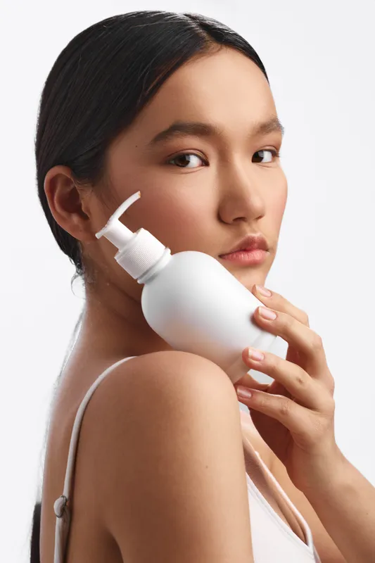 asian woman holds a lotion bottle near her shoulder in a clean studio, close up portrait with soft lighting, showcasing skincare and a calm, confident mood.