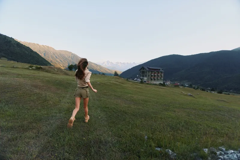 A woman in casual outdoorwear runs across a meadow towards a distant house, capturing a candid lifestyle moment of freedom, exploration, and connection with nature.