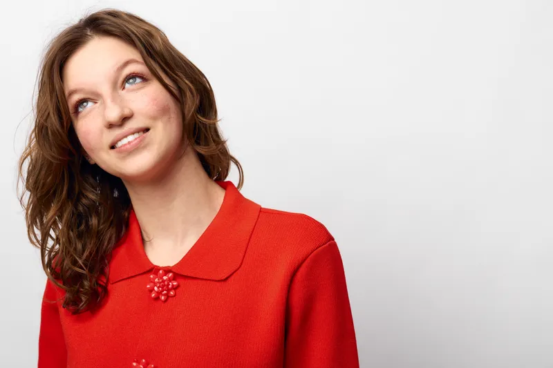 Young woman with curly brown hair wearing a red sweater looks up with a gentle smile. Studio portrait on white background. Concept of happiness, casual style, and youth.