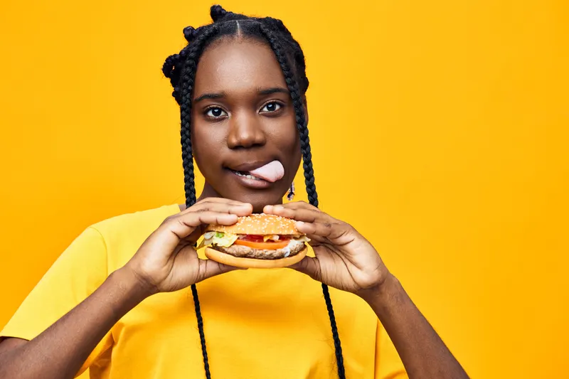 Young Black woman enjoying a colorful burger against a bright yellow background, showcasing joy and excitement with a playful expression