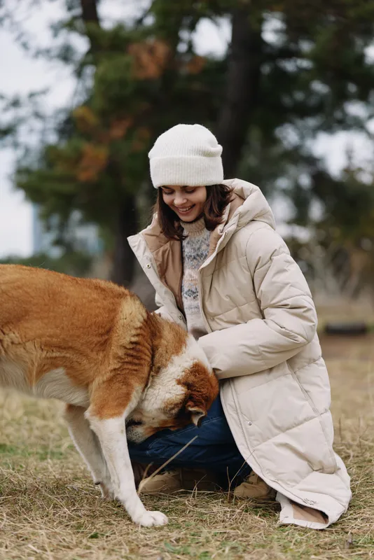 Dog, woman, outdoors, affection, interaction, winter, coat, cuddling with a canine companion on a chilly day in a park, wearing a warm jacket and beanie, capturing a tender moment of friendship and