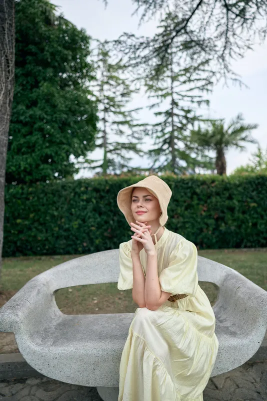 Woman wearing a hat sits on a curved stone bench in a sunny park, thinking with a gentle smile, creating a relaxing outdoor summer moment.