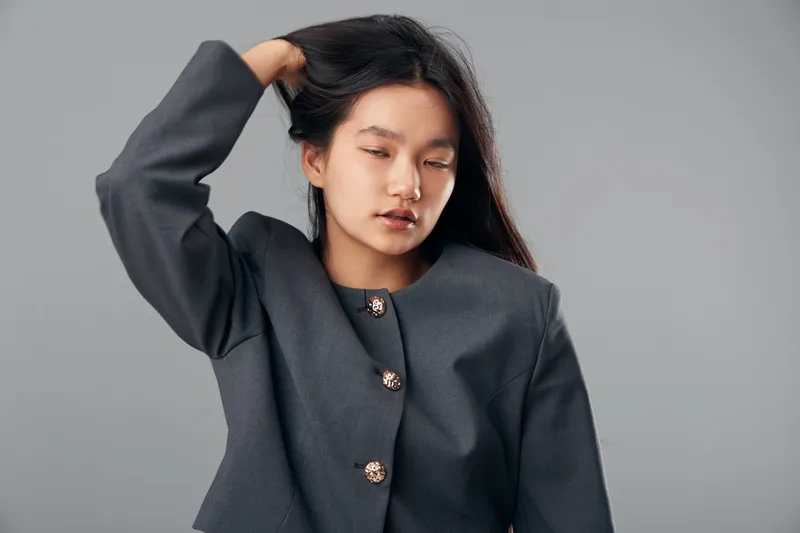 Young woman with dark straight hair posing with hand on head wearing a grey jacket with decorative buttons against plain grey background in studio portrait.