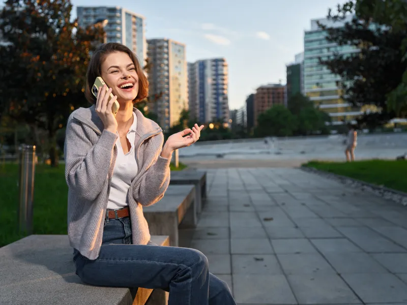 A cheerful young woman sits outdoors in a modern urban park, talking on a phone. She smiles brightly, projecting a positive and dynamic mood in a city setting.