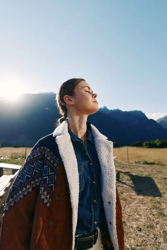 woman portrait outdoors with mountains and sunlight, wearing a jacket, serene profile with eyes closed. Rural field scenery and denim coat suggest travel, calm mood and natural beauty.