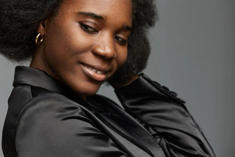 Elegant young African woman with natural hair, wearing a black satin jacket, smiling softly against a neutral gray background, embodies confidence and grace