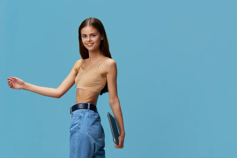 smiling woman with laptop wearing casual beige top and blue jeans stands against blue background with confident pose and friendly expression