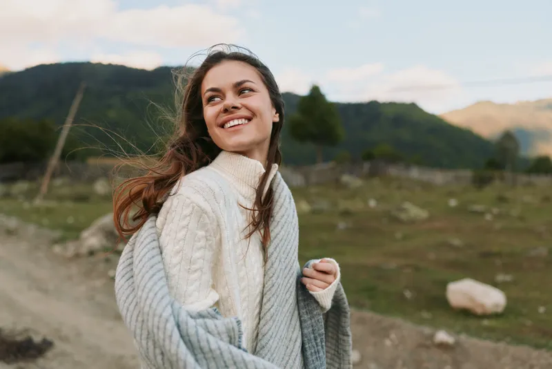 Smiling woman in a countryside landscape wears a chunky knit sweater, enjoying fresh air and open space, embodying casual outdoor lifestyle and wellbeing in the moment.