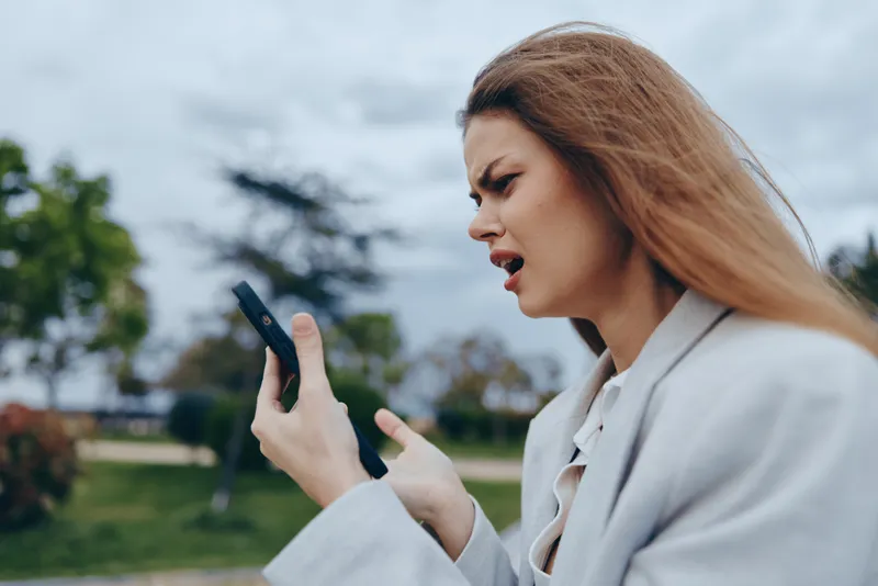 frustrated woman using smartphone outdoors, emotional upset young Caucasian woman with blonde hair in beige coat, cloudy park background, financial crisis concept