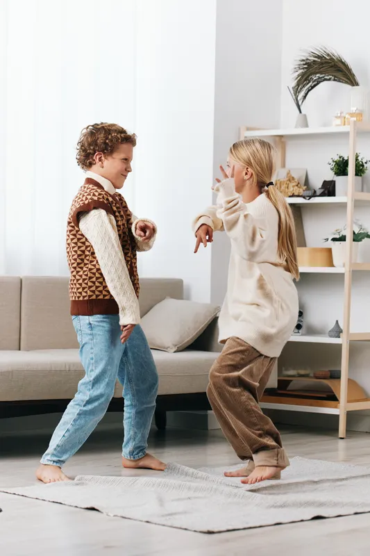 Two children dance playfully indoors, barefoot on a soft rug, wearing cozy sweaters and jeans. Lively expressions capture carefree rhythm and youthful joy.