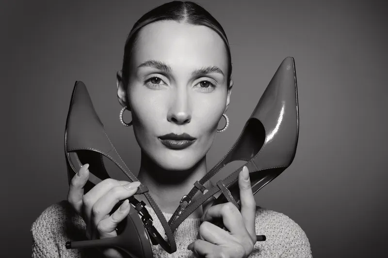 Portrait of a white young woman with fair skin holding high heels close to her face. Studio lighting highlights her facial features and earrings in black and white.