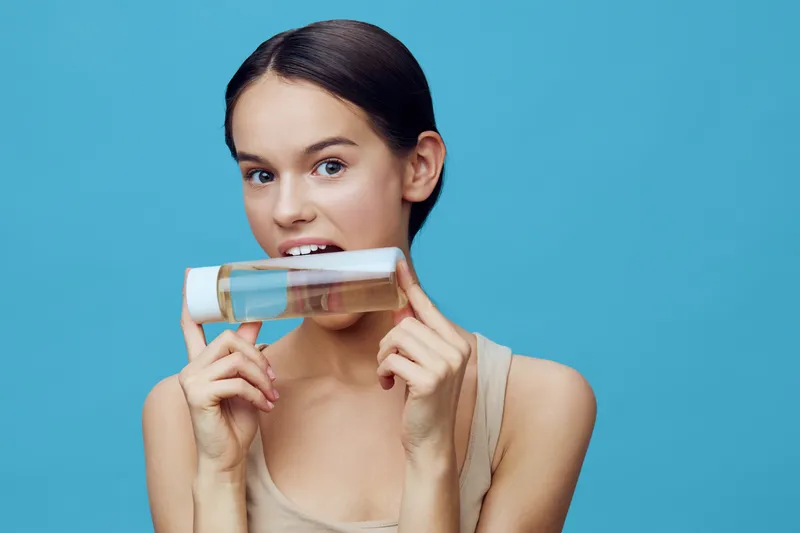 Young woman holding a skincare product, smiling playfully against a vibrant blue background, highlighting beauty and wellness concepts
