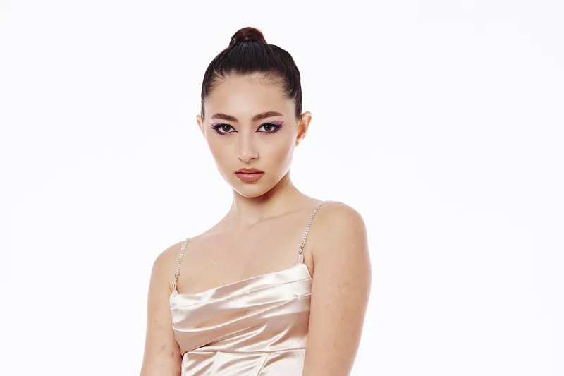 Young woman in a satin spaghetti strap dress poses confidently. Expressive makeup with purple eye shadow highlights her features. Studio shoot on white background.