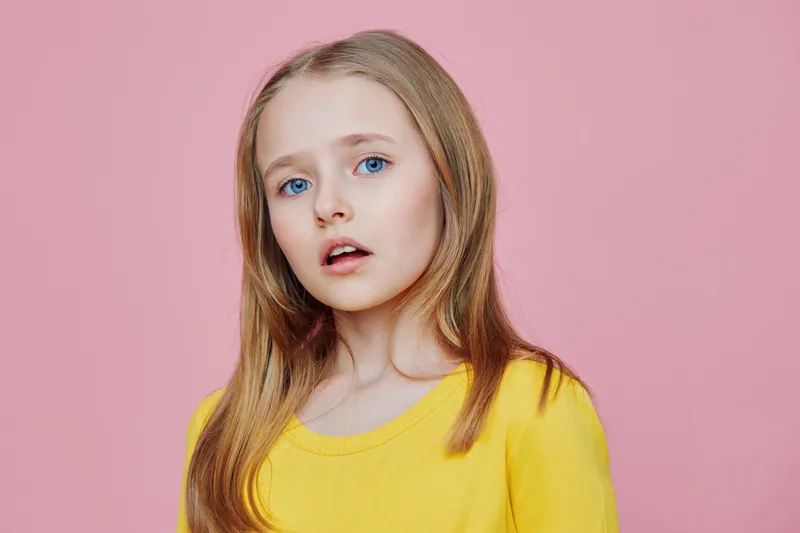 Portrait of a young girl with long blonde hair wearing a bright yellow sweater against a pink background, expressing surprise, ideal for concepts related to childhood and emotions