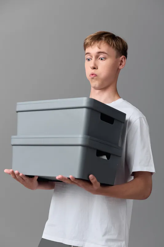 Young man in a white tee holding a stack of gray storage boxes, surprised expression, indoors, illustrating organization, packaging and stock handling in a simple workflow