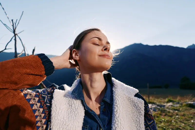 Woman portrait in sunlight on mountains, nature outdoors with relaxed smile and eyes closed. Relaxation moment in cozy jacket during hiking, calm golden hour and peaceful scenery.
