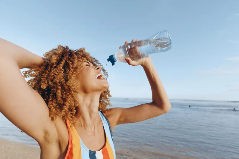 A fit woman on a sunny beach drinks water from a bottle, arms raised in celebration. The bright sea and sand backdrop highlights hydration, energy, and outdoor fitness.