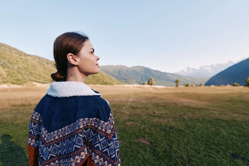 Woman portrait in a mountain landscape standing on a meadow, enjoying nature and outdoors. Traveler in a patterned jacket looks toward distant peaks across a wide grassy valley.
