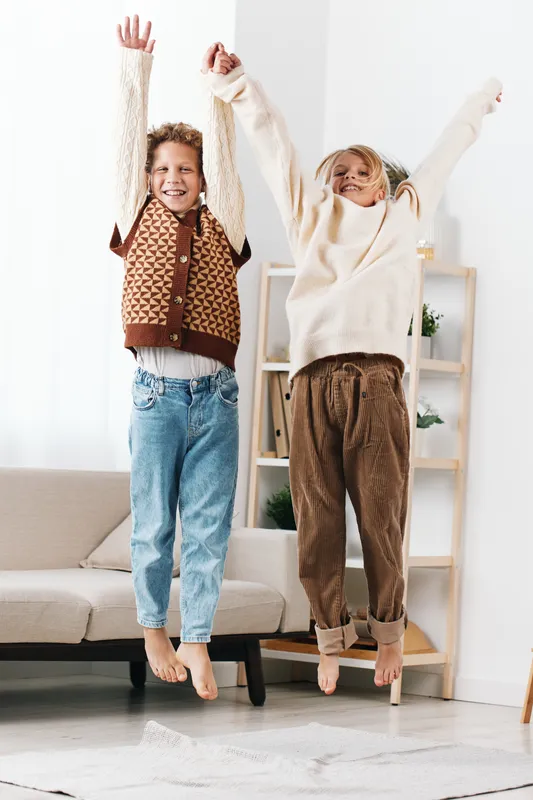Two joyful children jump with arms raised indoors, capturing a lively moment of energy, friendship, and carefree play in a bright home setting.