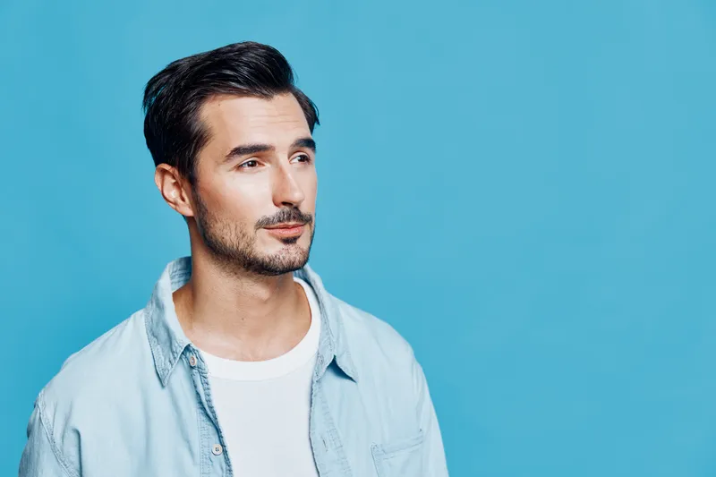 Thoughtful young man with stylish haircut wearing light blue shirt, looking to the side against a solid bright blue background. Portrait concept for lifestyle.