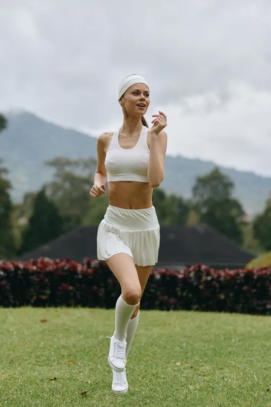 Fit young woman jogging in a white athletic outfit on a green field, surrounded by lush nature, conveying a healthy lifestyle and joy in exercise
